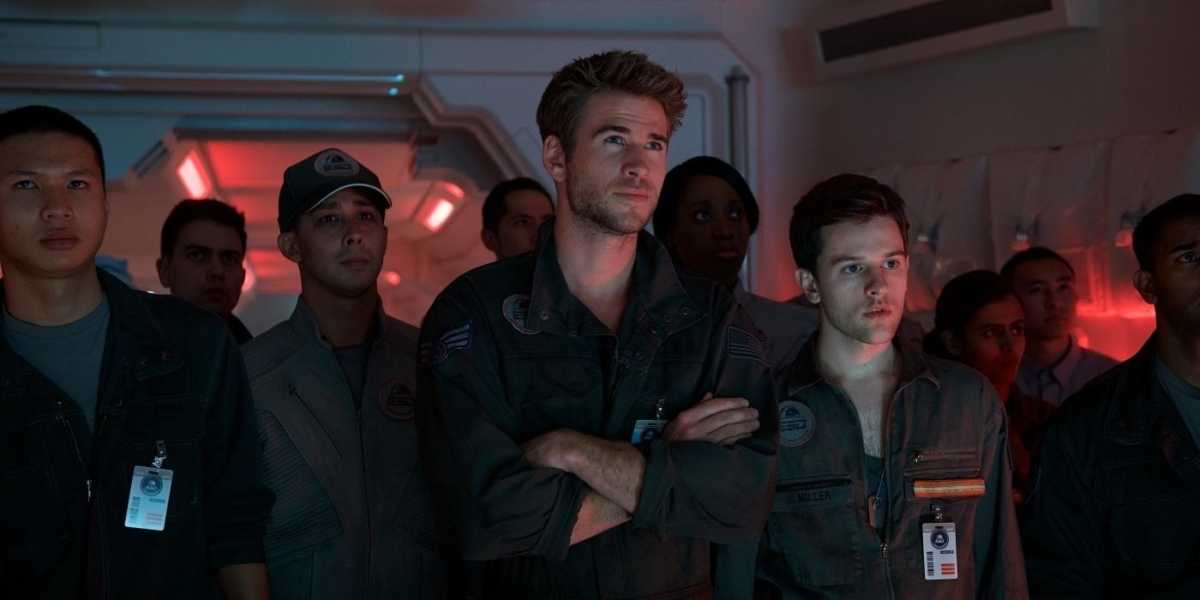 Homens e mulheres em trajes de voo estão em uma sala de controle iluminada por luzes vermelhas. Eles olham na mesma direção, com expressões sérias. O ator Liam Hemsworth está no centro, com os braços cruzados, e o ator Jessie Usher está à sua direita. Independence Day: O Ressurgimento, ficção científica, filme, Liam Hemsworth, Jessie Usher, sala de controle, trajes de voo.