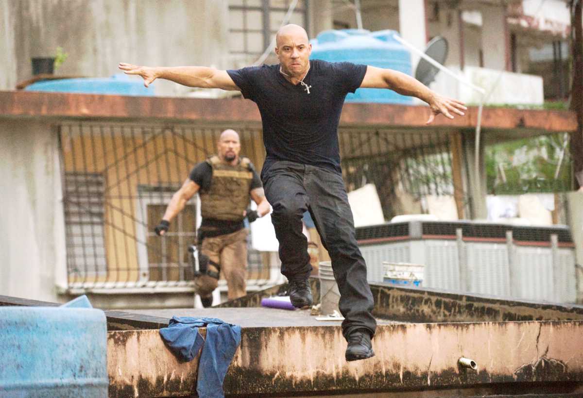 Vin Diesel como Dominic Toretto, em uma cena de perseguição no filme Velozes e Furiosos 5, pulando de um telhado com os braços abertos para se equilibrar. Ao fundo, o ator Dwayne "The Rock" Johnson, como o agente Luke Hobbs, o persegue. Vin Diesel usa uma camiseta preta e calças escuras, enquanto Dwayne Johnson usa uma camiseta cáqui e um colete tático.