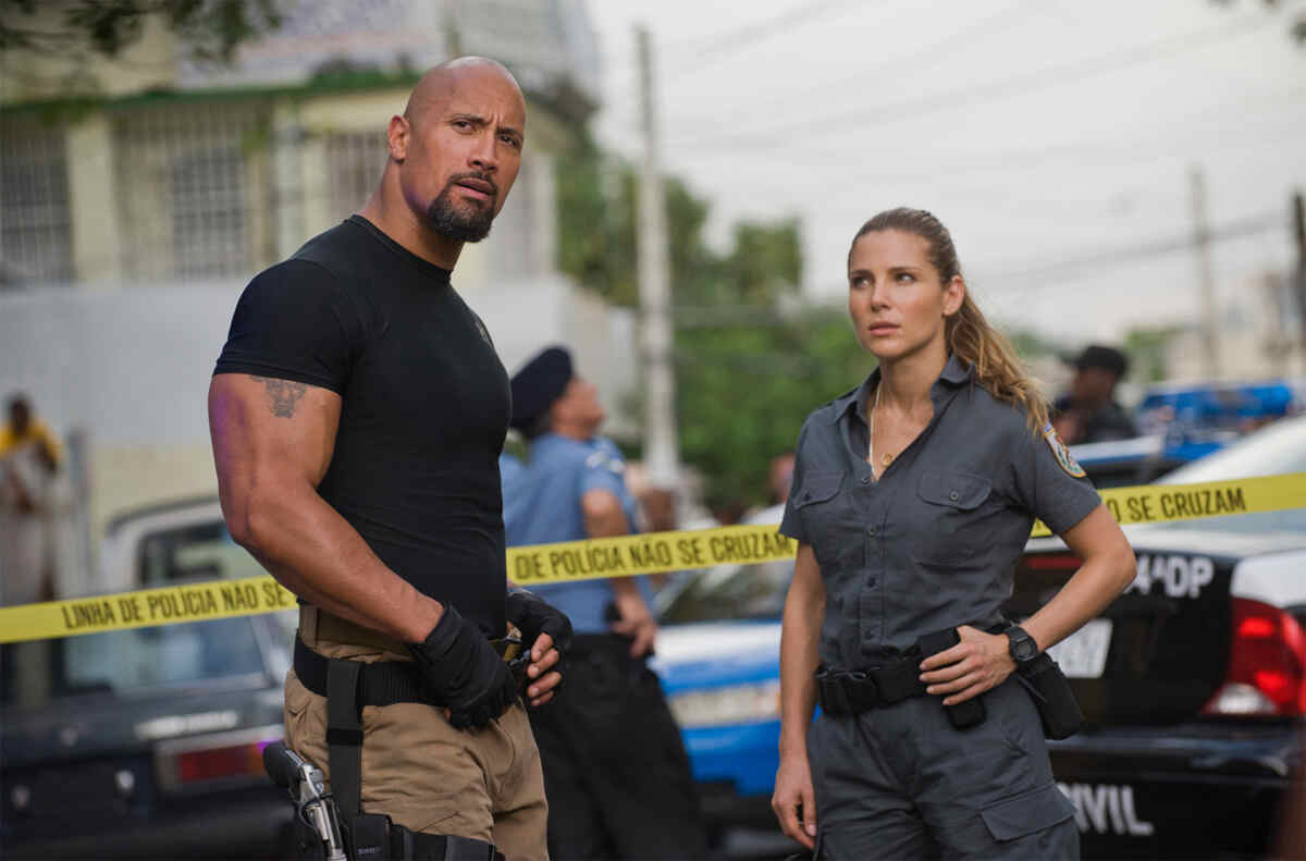 Dwayne "The Rock" Johnson como o agente Luke Hobbs e a atriz Elsa Pataky como a policial Elena Neves, parados e sérios em uma cena do filme Velozes e Furiosos 5. Eles estão em uma área isolada por uma fita amarela da polícia. Dwayne Johnson usa uma camiseta preta e calças cáqui, enquanto Elsa Pataky veste um uniforme de policial cinza
