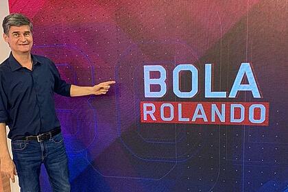 Fábio Piperno nos estúdios do Bola Rolando, no BandSports, em pé, apontando para o logo do programa que está ao seu lado