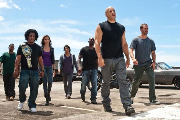 Um grupo de seis personagens do filme Velozes e Furiosos 5, liderado por Vin Diesel como Dominic Toretto e Paul Walker como Brian O'Conner, caminha em direção à câmera em uma cena ao ar livre. Eles estão em um estacionamento com um carro cinza antigo ao fundo. Vin Diesel usa uma regata preta, e Paul Walker usa uma camiseta cinza. Os outros personagens também estão com roupas casuais