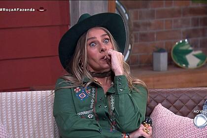 Adriane Galisteu, assustada e sentada no cenário de A Fazenda, com o dedo na boca e acompanhando o barraco na formação da roça