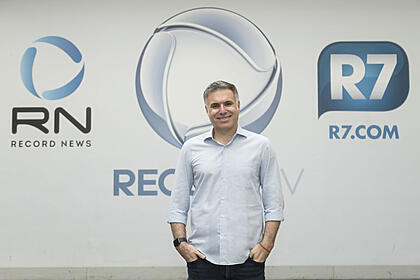 Narrador Marco de Vargas posando para foto em frente aos logos da Record TV, Record News e R7