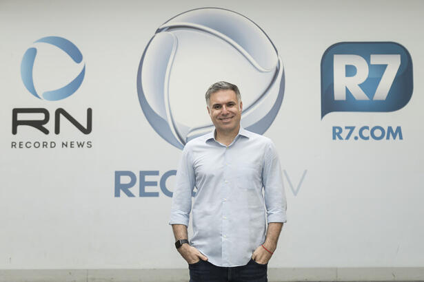 Narrador Marco de Vargas posando para foto em frente aos logos da Record TV, Record News e R7