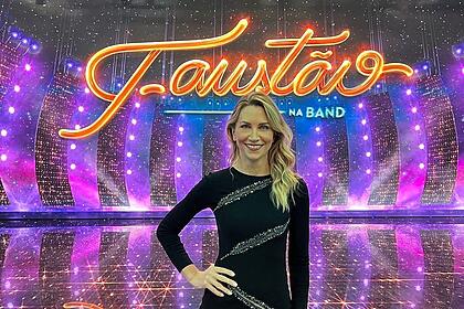Anne lottermann com um vestido preto, em frente ao telão nos estúdios do faustão na band, em uma foto posada