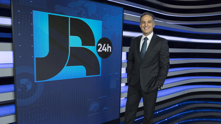 Leandro Stoliar de terno, parando em frente ao telão do JR 24h no estúdio e com as mãos nos bolsos