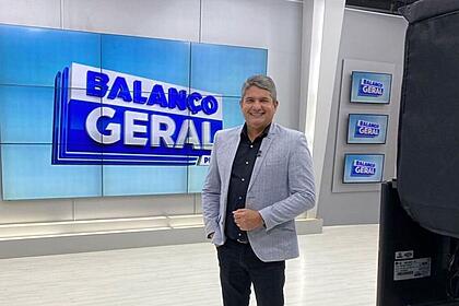Gernand lopes de terno cinza. Ele está sorrindo, em frente ao telão do balanço geral pe