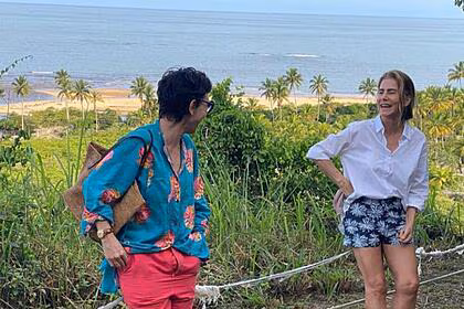 Adriana Calcanhotto aparece ao lao de Maitê Proença que sorrir. As duas estão em frente ao mar de Trancoso, Bahia