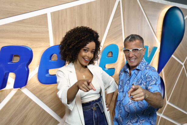 Alinne Prado e Eri Johnson, abraçados em frente ao telão com o logo da RedeTV! e apontando com o dedo para a câmera