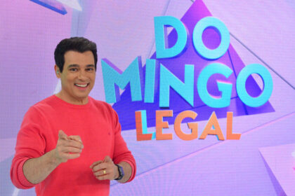 Celso Portiolli apontando para a câmera, em frente ao telão nos estúdios do Domingo Legal