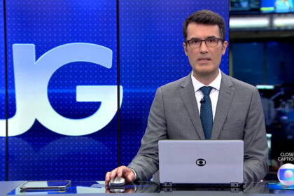 Fabio Turci sentado na bancada do Jornal da Globo, com o telão ao fundo e um notebook na bancada
