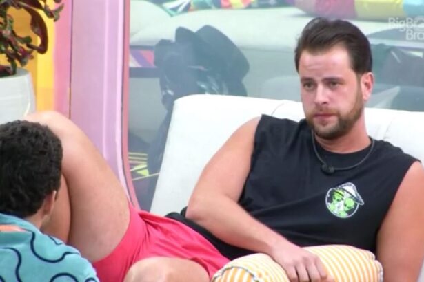 Gustavo sentado no sofá da área externa do BBB 22, vestido de uma camiseta preta olhando para frente conversando com Scooby.