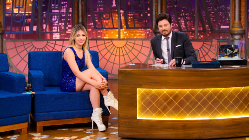 Melody de vestido azul e sentada nos estúdios do The Noite, ao lao de Danilo Gentili. Notas falsas de dinheiro estão no chão