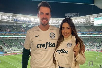 Gustavo e Laís abraçados, com a camisa do Palmeiras, tendo a visão do estádio do clube ao fundo