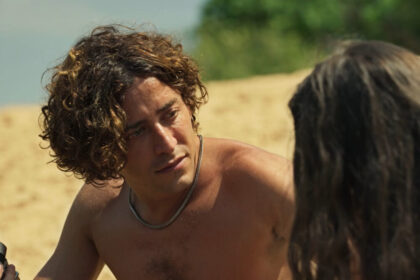 Jove (jesuíta barbosa) e juma (alanis guillen) em cena da novela pantanal. Ele com uma câmera fotográfica em mãos. Ela de costas aparecendo apenas os eu cabelo e a sombra