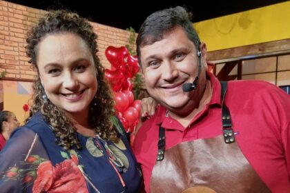 ApresentBianca Láua sorrindo ao lado do Padre Chef, ambos de vermelho e avental, na gravação do Sabor de Vida, da TV Aparecida