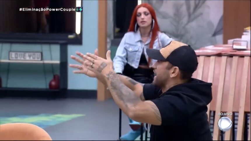 Hadballa de Boné com as mãos apontadas, atrás se encontra Brenda.