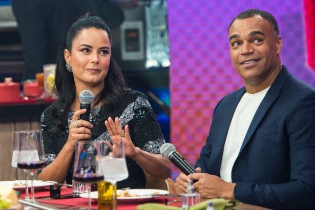 Luciele di Camargo de vestido preto, com o microfone na mão olhando para frente; Denilson de blazer azul e camisa básica branca com o microfone na mão olhando pra cima na gravação do Faustão da Band, da Band