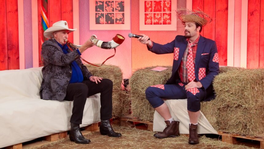 Sergio Reis sentado, de chapéu de cowboy, jaqueta e camisa azul, calça e sapatos pretos tocando berrante ao lado de Danilo Gentili, que está caracterizado com roupa junina, chapéu, segurando o microfone na ponta do berrante, durante gravação do The Noite, do SBT