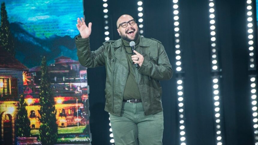 Tiago Abravanel de jaqueta, camisa e calça jeans cinza, com microfone na mão, acenando para a plateia durante gravação do Faustão na Band