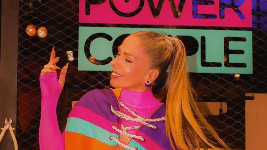 Adriane Galisteu no cenário do Power Couple Brasil apontando para o logo do programa