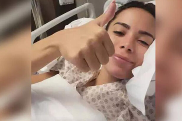 Anitta faz o sinal de positivo com a mão, cantora está deitada em um leito de hispital