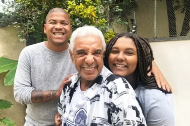 Damião de camisa cinza, segurando no ombro de Agnaldo Timoteo, que está de camisa branca estampada com detalhes pretos, sorrindo, ao lado da filha, Keyty, que está sorrindo, com penteado de tranças e camisa cinza