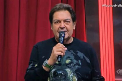 Fausto Silva com microfone na mão direita, de camisa preta estampada com um cachorro durante apresentação do Faustão na Band