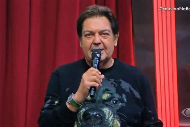 Fausto Silva com microfone na mão direita, de camisa preta estampada com um cachorro durante apresentação do Faustão na Band