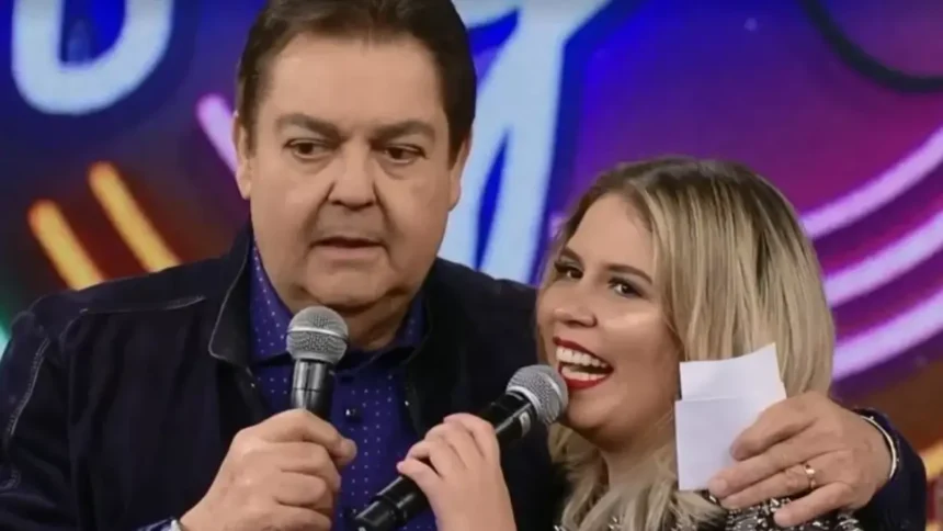 Faustão de camisa preta, segurando o microfone com a mão direita, abraçando Marília, que está de vestido preto, sorrindo, com o microfone próximo a boca, durante gravação do Domingão do Faustão, da TV Globo
