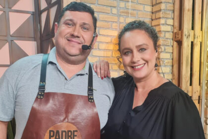 Padre Chef Evandro de camisa social cinza e avental marrom com a logo do programa Sabor de Vida e Bianca Lauá de camisa social preta, colar de ouro, sorrindo, com a mão no ombro do Chef durante gravação de Sabor de Vida, da TV Aparecida
