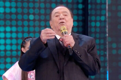 Raul Gil de palitó preto, segurando o microfone dourado enquanto grava seu programa no SBT