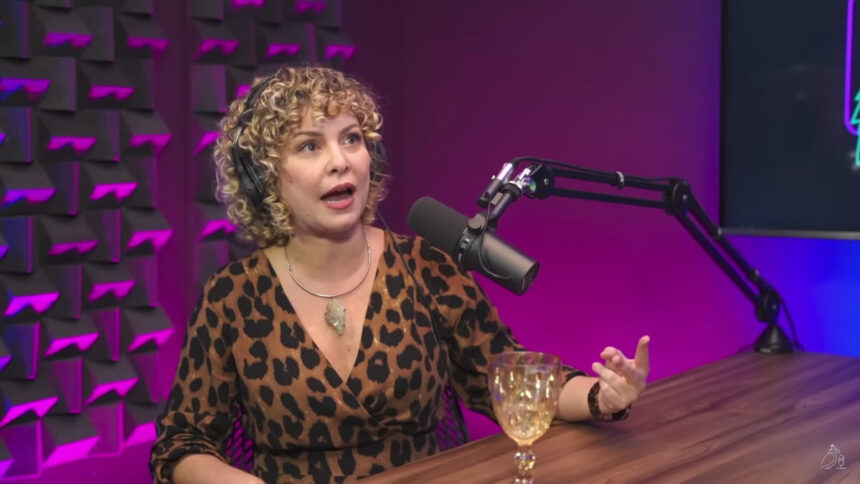 Bárbara Borges de vestido animal print de onça, decotada, colar de prata sentada numa cadeira de ferro preta, com a mão na bancada do podcast Papagaio Falante