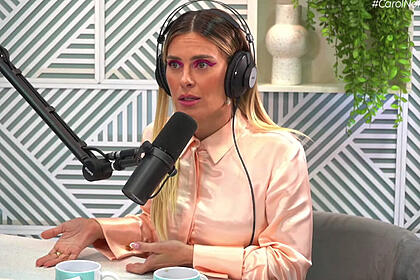 Carolina Dieckman de camisa social rosa claro, fone de ouvido, sentada, próxima ao microfone durante entrevista ao Poccast