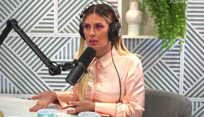 Carolina Dieckman de camisa social rosa claro, fone de ouvido, sentada, próxima ao microfone durante entrevista ao Poccast