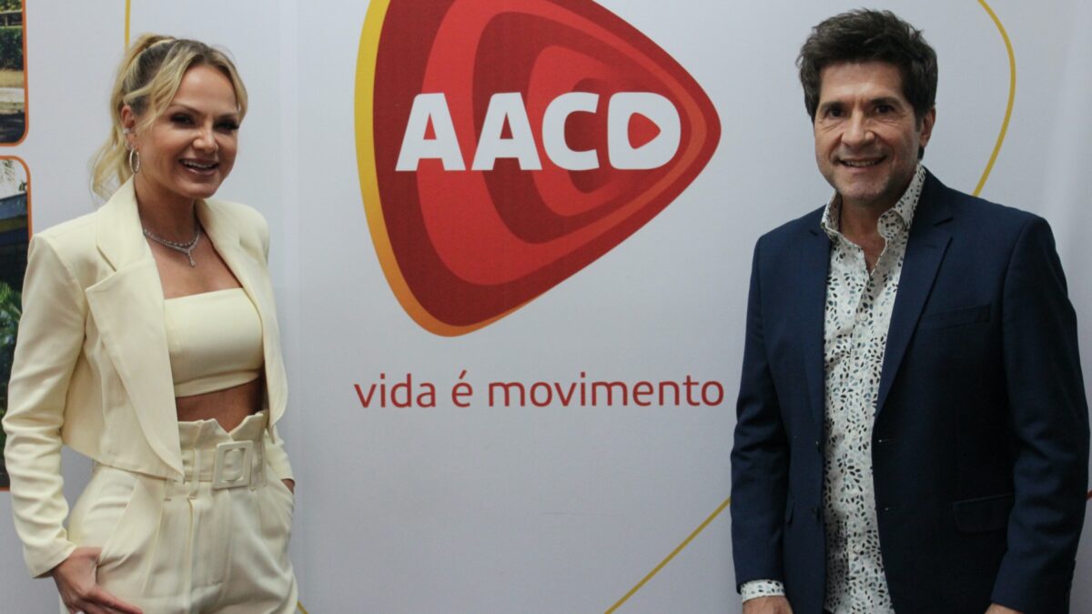 Eliana com blazer e calça bege claro, com a mão no bolso, sorrindo ao lado da logo do AACD; Daniel de blazer e calça azul e camisa social, sorrindo