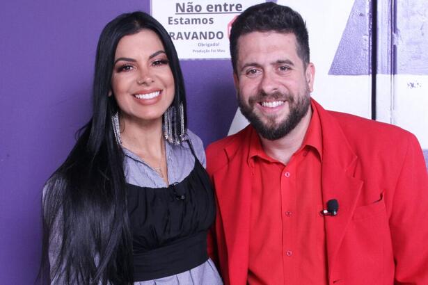 Evelyn Regly com top preto, camisa social roxa, sorrindo, cabelo preto solto ao lado de Mauricio Meireles, de blazer, camisa e calça vermelha, sorrindo, durante gravação do Foi Mau, da RedeTV!