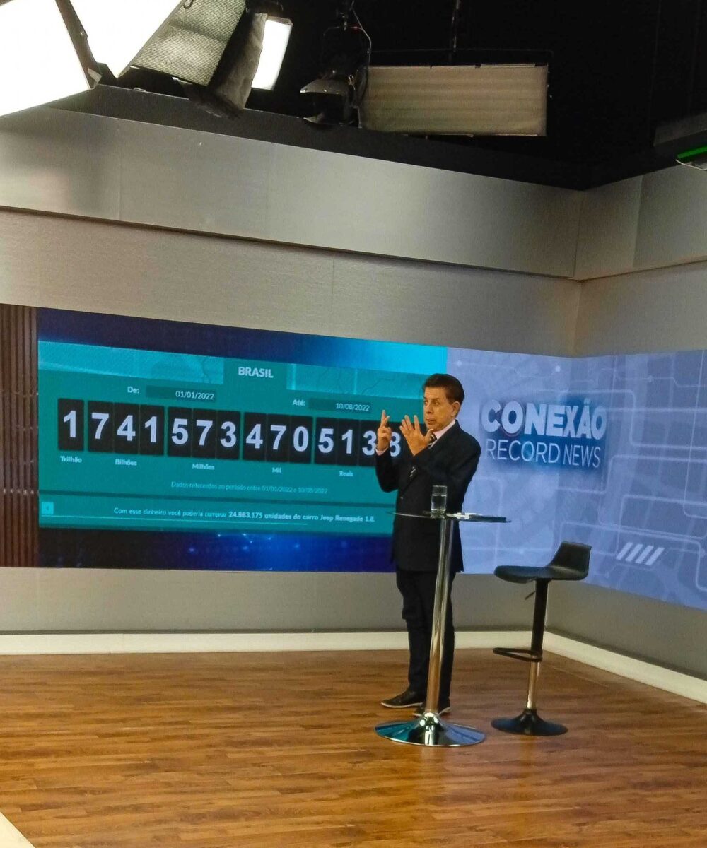 Heródoto Barbeiro em trecho do Conexão Record News, com o telão ao fundo. A imagem também contém os bastidores do chão do cenário do teto com os spots de iluminação