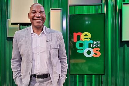 Apresentador João Vicente de terno, com a mão no bolso, nos estúdios do programa Negros em Foco, em frente a um telão