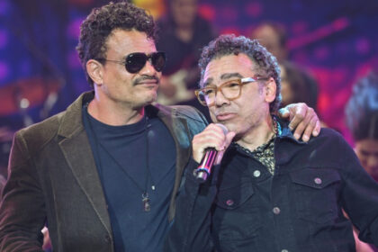 Leo Maia de óculos de sol preto, blazer preto e camisa azul marinho com a mão esquerna no ombro de Simoninha; Simoninha de camisa manga longa preta segurando o microfone preto com a mão direita durante gravação do Faustão na Band