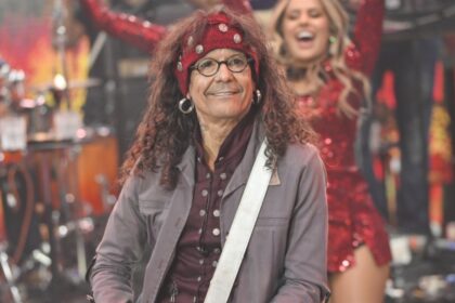 Cantor Luiz Caldas com blazer cinza, camisa preta e vermelha estampada e bandana na cabeça, tocando baixo, sorrindo durante performance do Faustão na Band