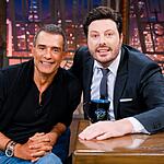 Marcos Pasquim sorrindo, de camisa preta e calça jeans, com as mãos na coxa, sentado no sofá azul ao lado de Danilo Gentili durante gravação do The Noite, do SBT