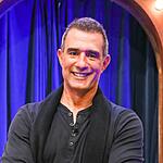 Marcos Pasquim de camisa social preta, sorrindo, com os braços cruzados durante gravação do The Noite, do SBT