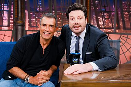 Marcos Pasquim sorrindo, de camisa preta e calça jeans, com as mãos na coxa, sentado no sofá azul ao lado de Danilo Gentili durante gravação do The Noite, do SBT