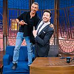 Marcos Pasquim sorrindo com camisa social preta e calça jeans azul em cima do sofá azul ao lado de Danilo Gentili durante gravação do The Noite, do SBT