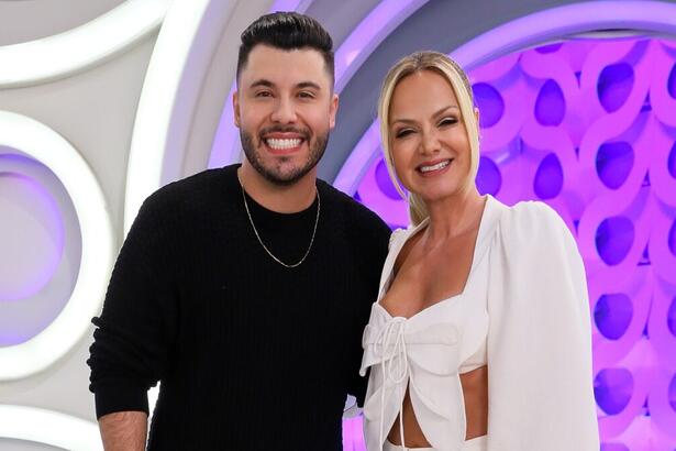Murilo Huff de camisa preta, colar de prata, sorrindo, ao lado de Eliana, com croppet branco e calça branca durante gravação do Programa Eliana, do SBT