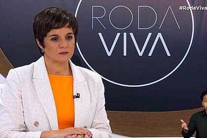 Vera Magalhães de blazer branco, camisa cinza, microfone preto grampeado no blazer, com semblante sério, sentada num banco durante gravação do Roda Viva, da TV Cultura