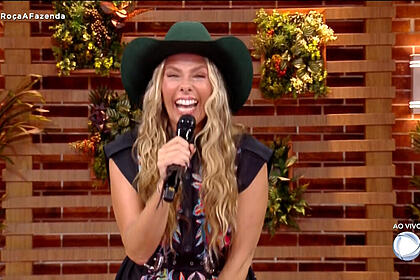 Adriane galisteu, com chapéu na cabeça, microfone na mão e sorriso aberto