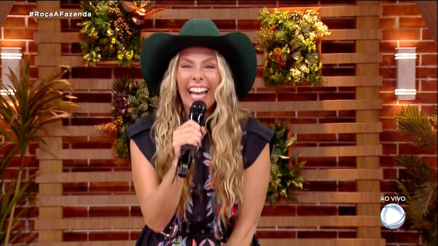Adriane Galisteu, com chapéu na cabeça, microfone na mão e sorriso aberto