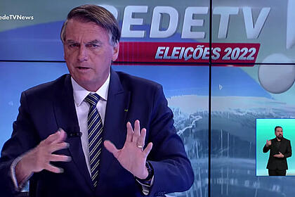 Bolsonaro de palitó, preto, gravata listrada preta a branca, camisa social branca, gesticulando com as mãos, sentado durante entrevista na RedeTV!